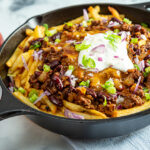 A cast-iron skillet of chili cheese fries topped with sour cream and chopped onions