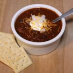 A bowl of chili with a dollop of sour cream, onions, and cheese on top and a few crackers next to it